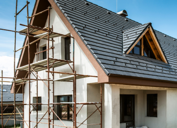 Zweistöckiges Haus im Rohbau mit steil geneigtem Dach und dunkelgrauen Ziegeln. Links am Gebäude ist ein Gerüst aufgebaut. Die Fassade ist weiß, und auf beiden Etagen sind mehrere Fenster zu sehen. Eine kleine Gaube befindet sich im Dach. Der Bereich um das Haus ist noch nicht fertiggestellt, mit sichtbaren Gras- und Erdflächen.