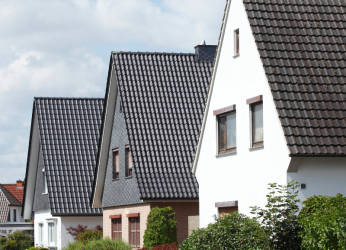 Drei nebeneinander stehende Häuser mit dunklen, steil geneigten Ziegeldächern. Das erste und dritte Haus haben eine weiße Fassade, das mittlere Haus kombiniert weiße Fassade mit roten Ziegeln. Vor den Häusern befinden sich Büsche und Grünpflanzen. Der Himmel ist teilweise bewölkt. Es handelt sich um Wohngebäude.