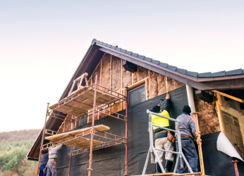 Baustelle mit einem Haus, dessen Außenwände teilweise mit schwarzer Schutzverkleidung versehen sind. Arbeiter befestigen Dämm- und Wetterschutzmaterialien mithilfe von Werkzeugen. Ein Gerüst umgibt das Gebäude, um Zugang zu höheren Bereichen zu ermöglichen. Im Hintergrund sind Bäume und Hügel zu sehen.