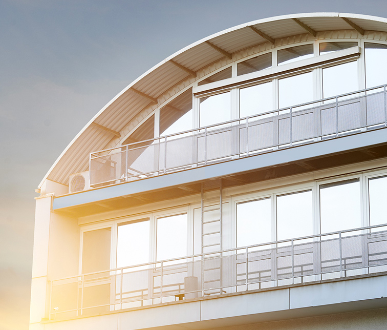 Oberer Teil eines modernen Gebäudes mit geschwungenem Dach, großen Fenstern und Balkonen mit Metallgeländern. Auf einem Balkon befindet sich eine Klimaanlage. Sonnenlicht beleuchtet die Fassade warm